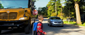 A young boy walks in front of a stopped school bus and car while the stop arm is out and blinking so he can cross safely