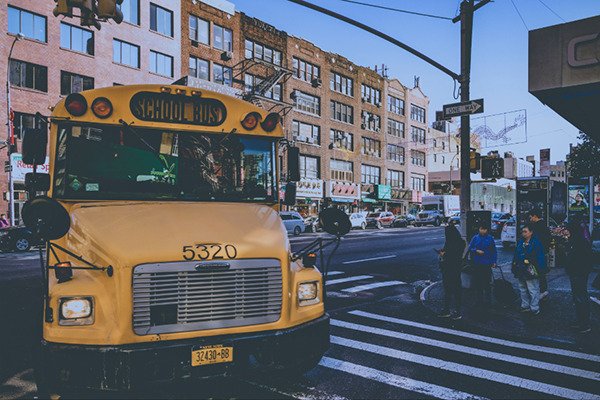 A school bus turning the corner of a busy street during a bright sunny day. Pedestrians wait for the bus to go in order to cross safely.